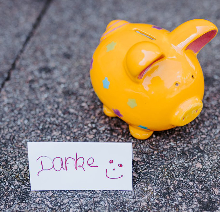 Ein orangenes Sparschwein aus Keramik steht auf einem Steinboden. Davor steht ein Papierschild, auf dem mit Handschrift Danke und ein Smiley geschrieben sind.