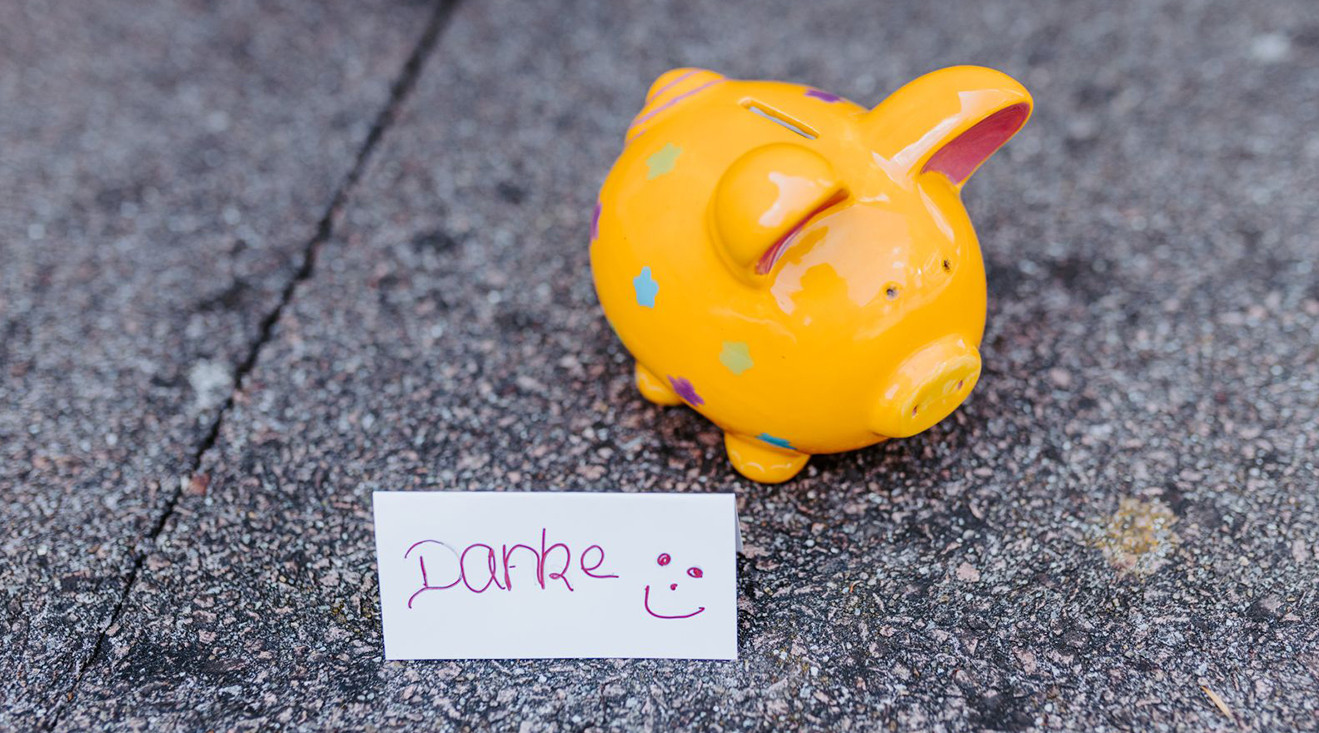 Ein orangenes Sparschwein aus Keramik steht auf einem Steinboden. Davor steht ein Papierschild, auf dem mit Handschrift Danke und ein Smiley geschrieben sind.