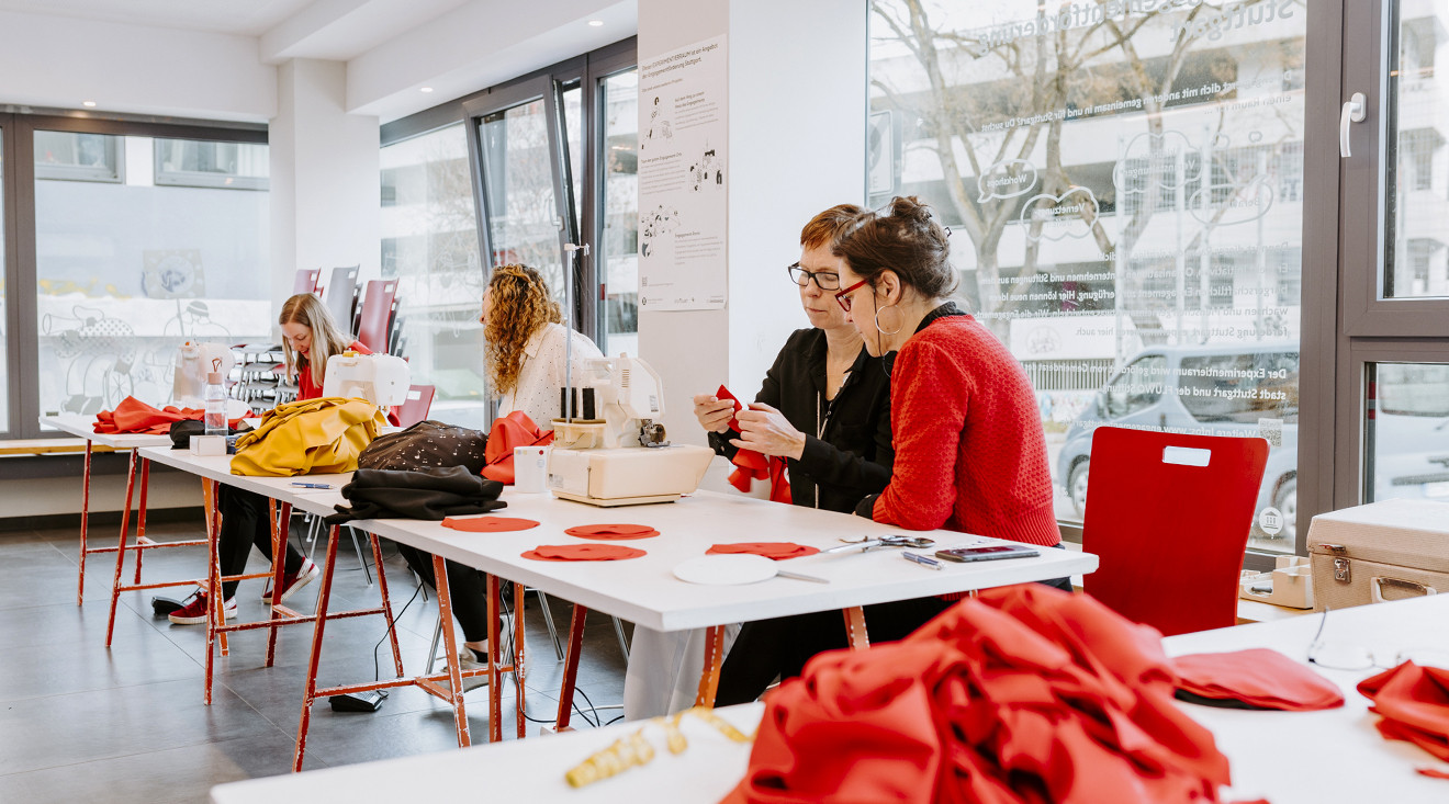 Vier Frauen sitzen an Tischen und nähen mit Nähmaschinen. Eine Frau beugt sich zu einer anderen und erklärt ihr etwas. 