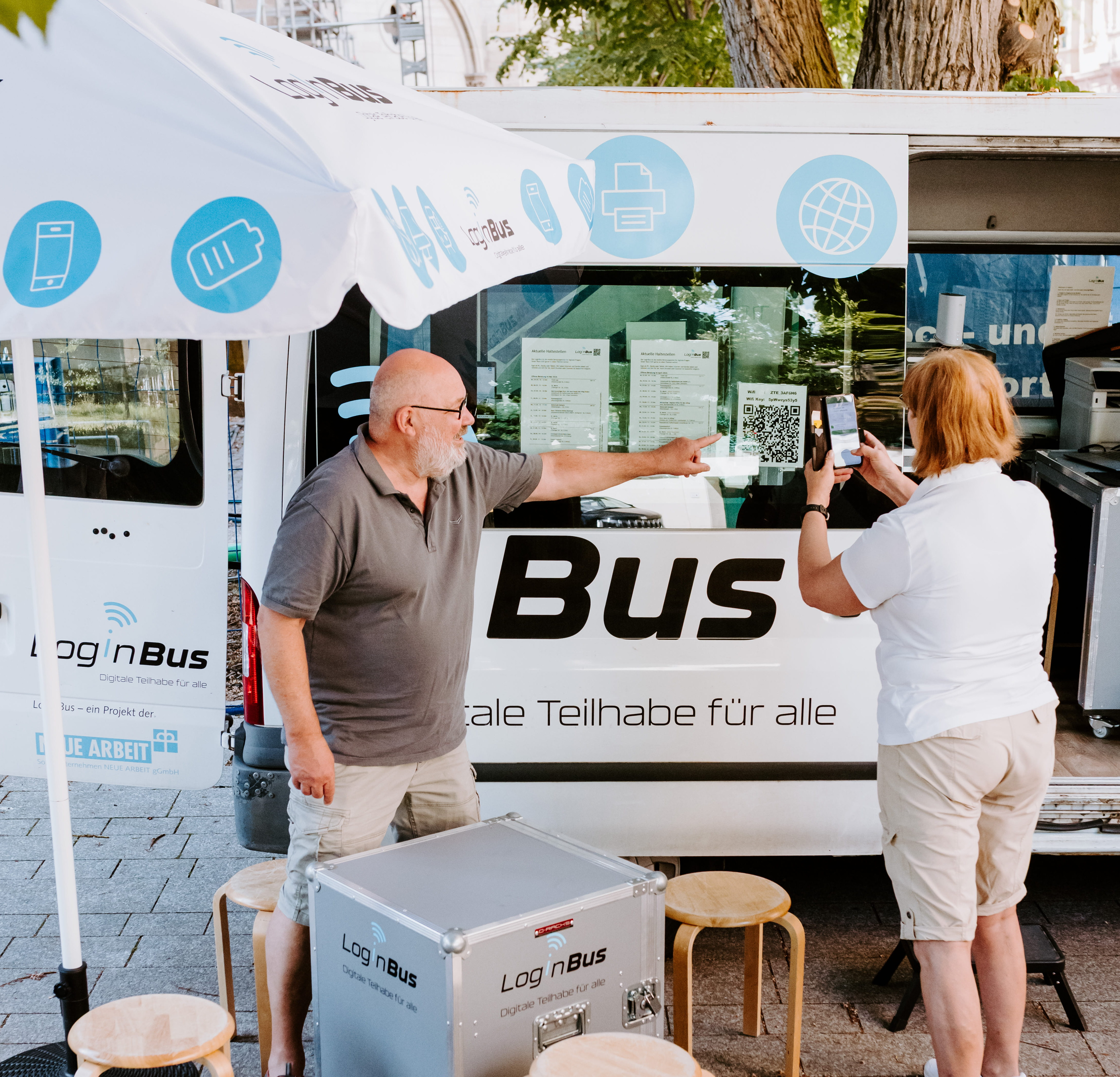 Eine Frau und ein Mann stehen vor einem Kleinbus. Der Mann zeigt auf ein Plakat, das am Bus befestigt ist. Die Frau macht ein Foto davon. 
