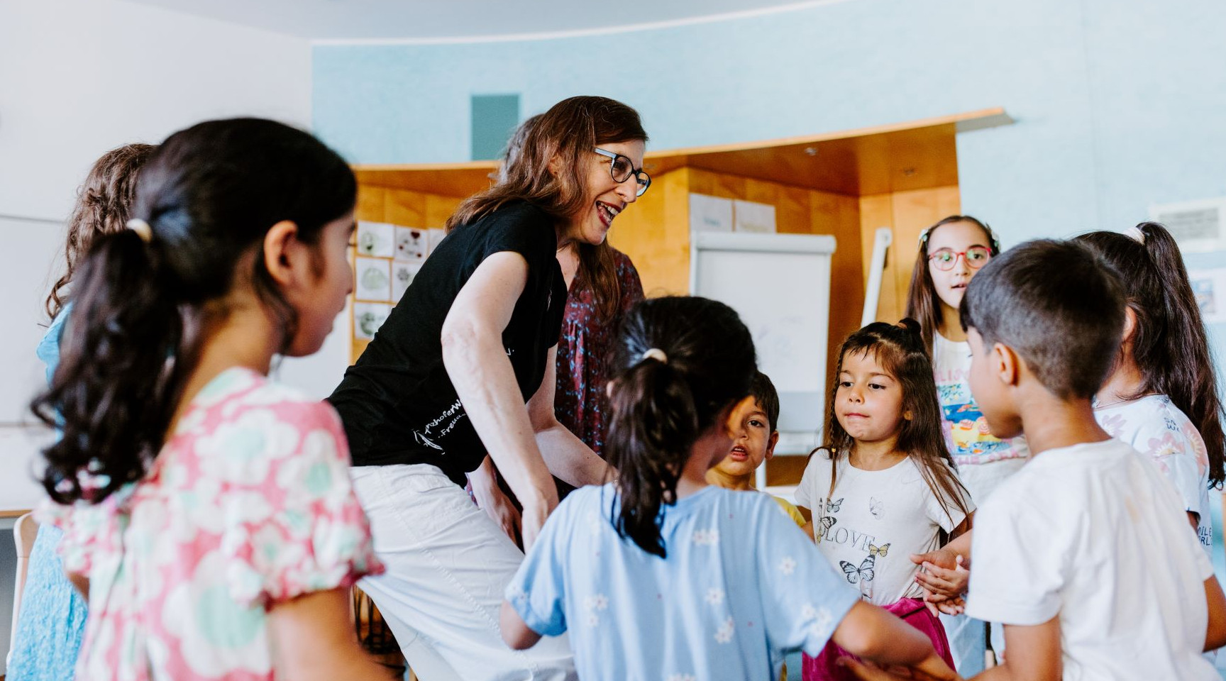 Eine Frau steht inmitten einer Gruppe von Kindern, die sich an den Händen halten. Sie tanzen. 