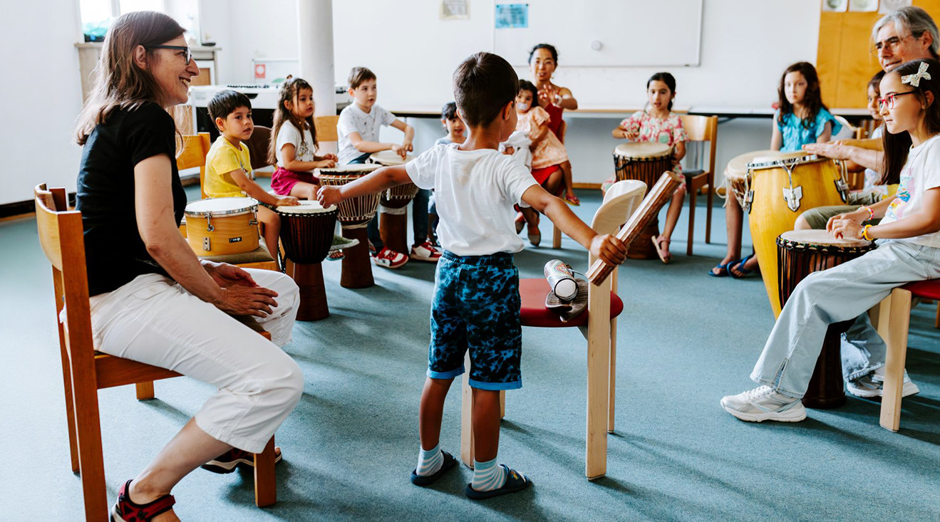 Eine Gruppe von Kindern und Erwachsenen sitzt im Kreis. Jedes Kind und jeder Erwachsene spielt eine Trommel. Ein Kind steht vor der Gruppe und dirigiert. 
