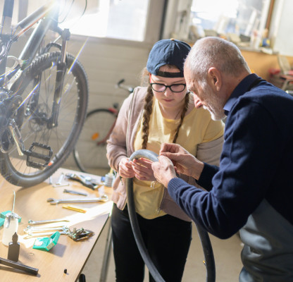 Eine junge Frau und ein älterer Mann arbeiten gemeinsam an einer Werkbank und beugen sich über einen Fahrradschlauch. Im Hintergrund steht ein Fahrrad.