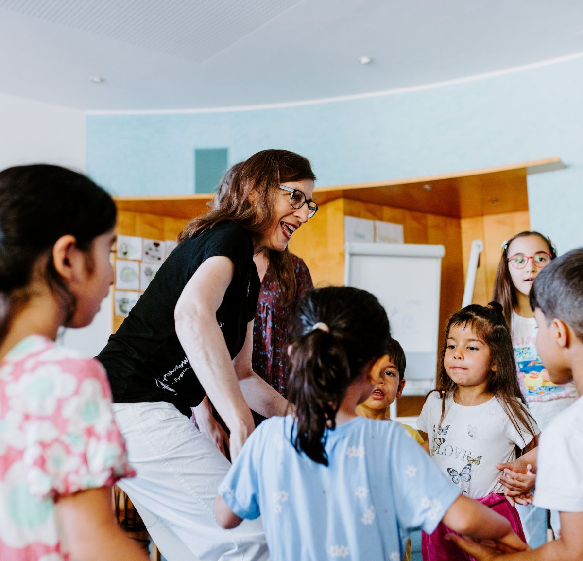 Eine Gruppe von Kindern hält sich an den Händen. Eine Frau steht in der Mitte und lacht. Sie tanzen. 