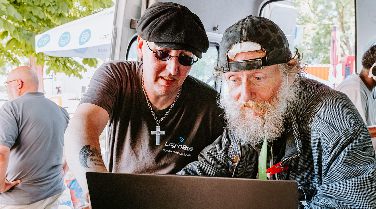 Zwei Männer, einer im mittleren Alter, einer im höheren Alter schauen gemeinsam auf einen Laptop. Der jüngere Mann zeigt auf den Bildschirm. 