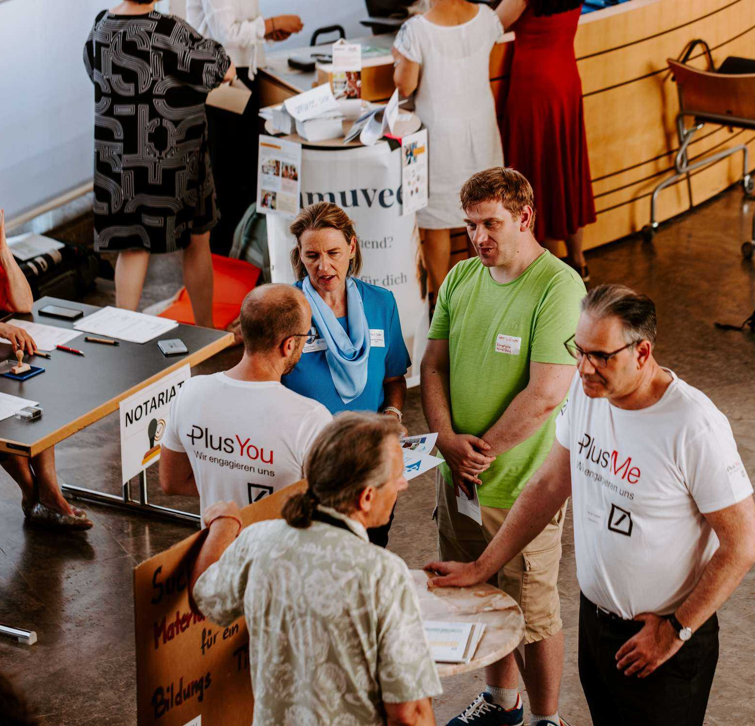 Drei Besucher einer Messe unterhalten sich mit zwei Betreuern eines Info-Stehtisches. Im Hintergrund sind weitere Besucher und Stände zu sehen.