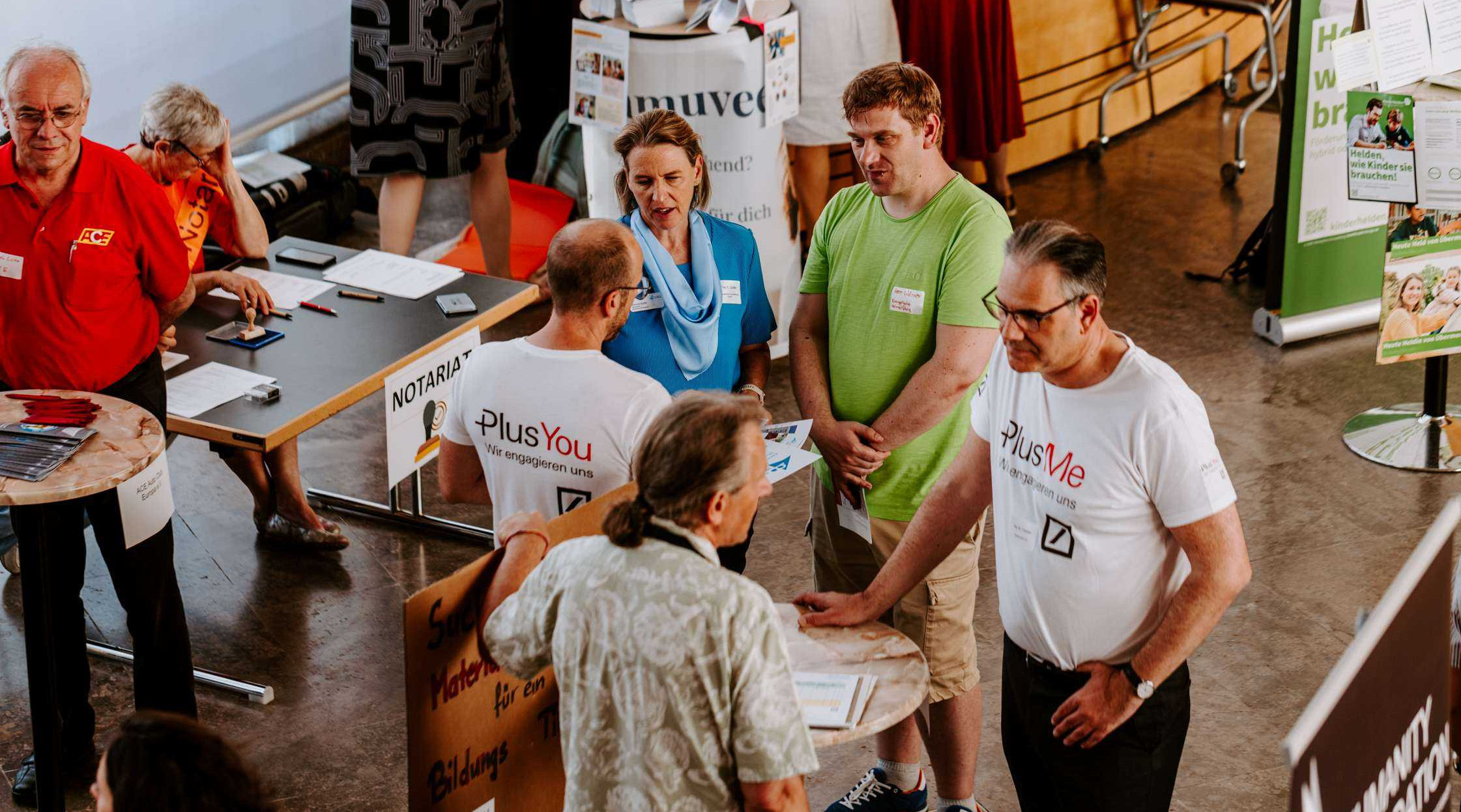 Drei Besucher einer Messe unterhalten sich mit zwei Betreuern eines Info-Stehtisches. Im Hintergrund sind weitere Besucher und Stände zu sehen.