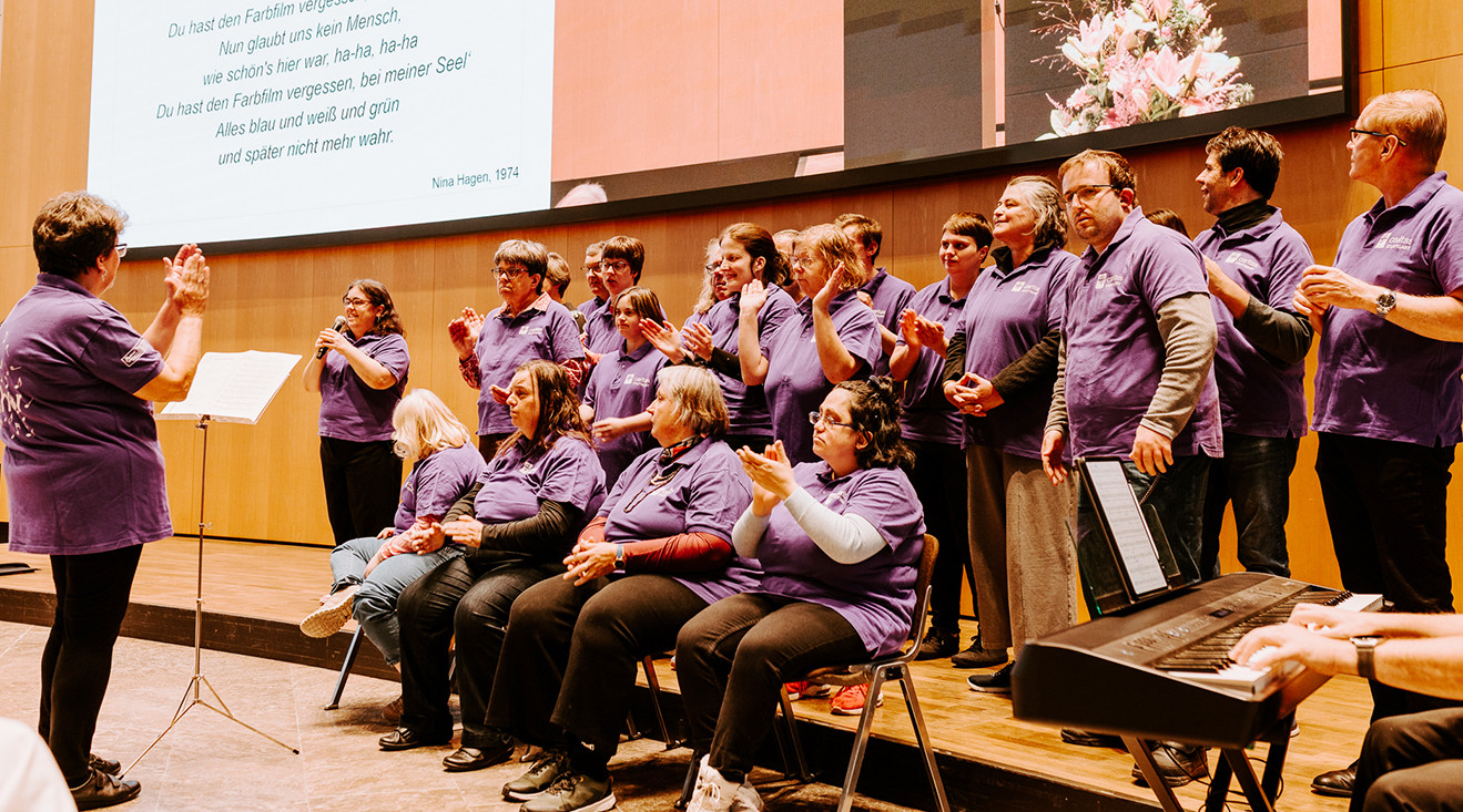 Ein Chor singt auf einer Bühne. Alle tragen lila T-Shirts. Einige klatschen. Davor steht die Chorleiterin und dirigiert. Auf eine Leinwand im Hintergrund ist der Liedtext projiziert: Du hast den Farbfilm vergessen. 