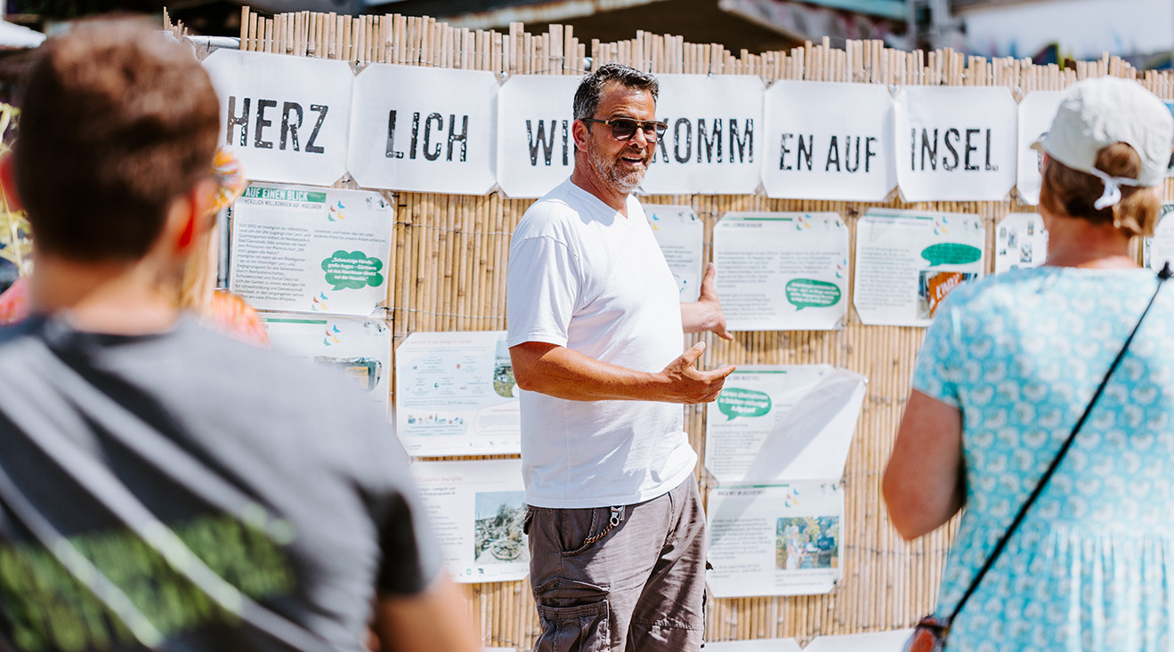 Ein Mann mit Sonnenbrille steht vor einer Bretterwand, an der Plakate angebracht sind und erklärt zwei Personen etwas, die nur von hinten zu sehen sind.