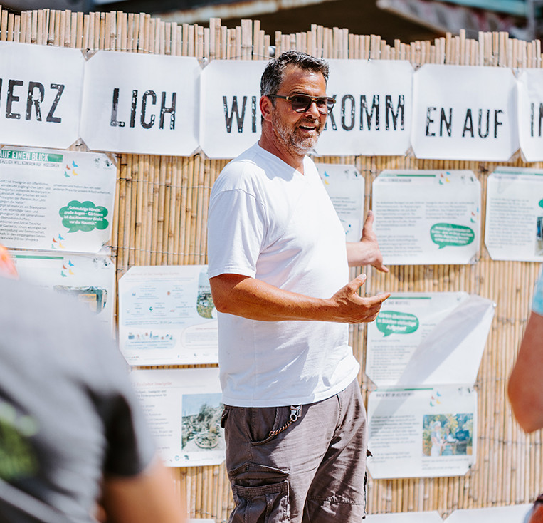 Ein Mann mit Sonnenbrille steht vor einer Bretterwand, an der Plakate angebracht sind und erklärt zwei Personen etwas, die nur von hinten zu sehen sind.
